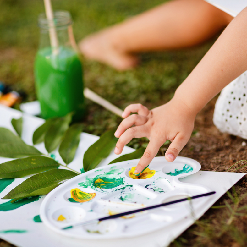 Child's hand playing with watercolor paint palette, sitting outside on green grass with a green beverage in background