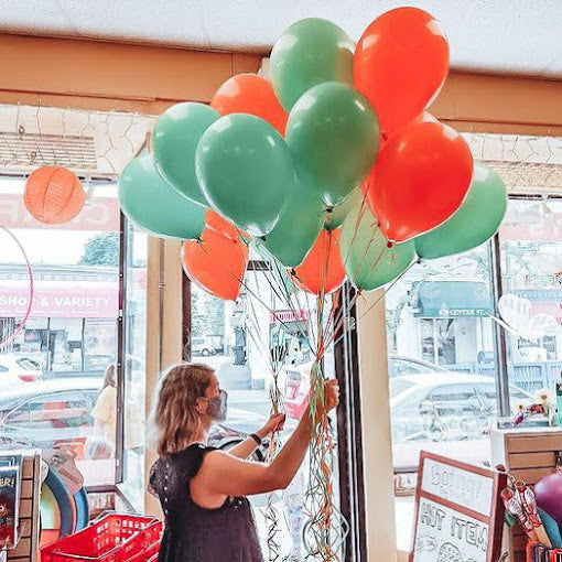 Person with light skin and hair wearing face mask and sleeveless top holds a bunch of light blue and red helium balloons, with the background a well-lit view out to the street from store windows. 