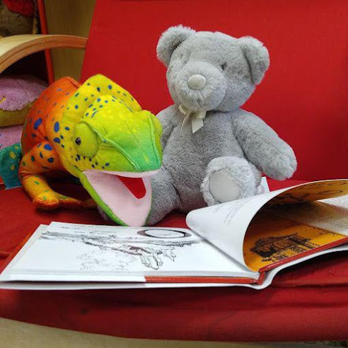 A gray teddy and a bright orange, yellow, and green chameleon puppet sit in a red chair witha picture book open in front of them